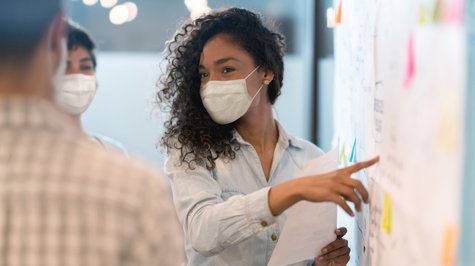 young woman wearing mask talking and pointing at a wall